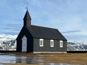 Schwarze Kirche mit weißen Details vor schneebedeckten Bergen und blauem Himmel in Island