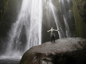 Person steht mit ausgestreckten Armen auf einem großen Felsen vor einem mächtigen Wasserfall an moosbedeckten Klippen in Island