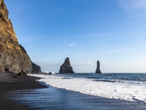 Schwarzer Sandstrand mit hoher Klippe, brechenden Wellen und zwei Felsnadeln im Meer unter blauem Himmel in Island