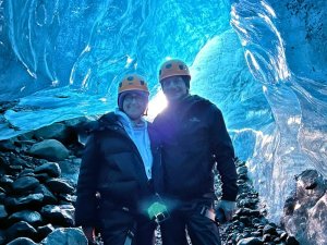 Zwei Menschen mit Helmen stehen in einer leuchtend blauen Eishöhle mit felsigem Boden in Island