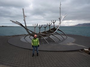 Frau in grüner Jacke und rosa Mütze steht neben der stählernen Wikinger-Skulptur Sólfar am Meer an einem bewölkten Tag in Reykjavík, Island