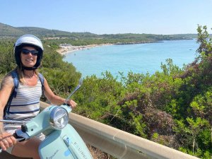 Reisende auf einer Vespa vor strahlend blauen Wasser in Alghero, Italien