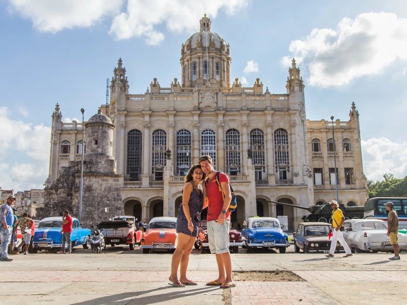 Lächelndes Paar vor einem großen Kuppelgebäude mit Oldtimern und Panzer auf der Straße in Havanna, Kuba