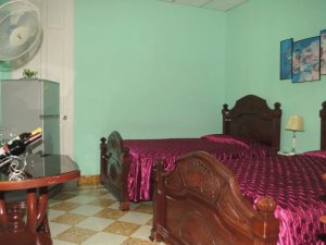 Mintgrünes Zimmer mit zwei Betten mit lila Tagesdecken, Minikühlschrank, Ventilator und schwarz-weißem Fliesenboden in Baracoa, Kuba