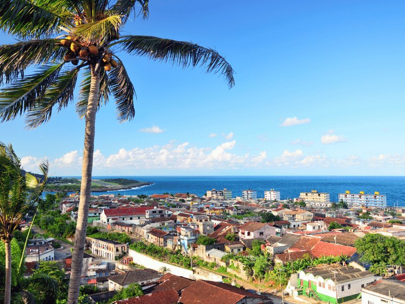 Blick über eine Küstenstadt mit Palme im Vordergrund und dem Ozean unter blauem Himmel in Santiago de Cuba, Kuba