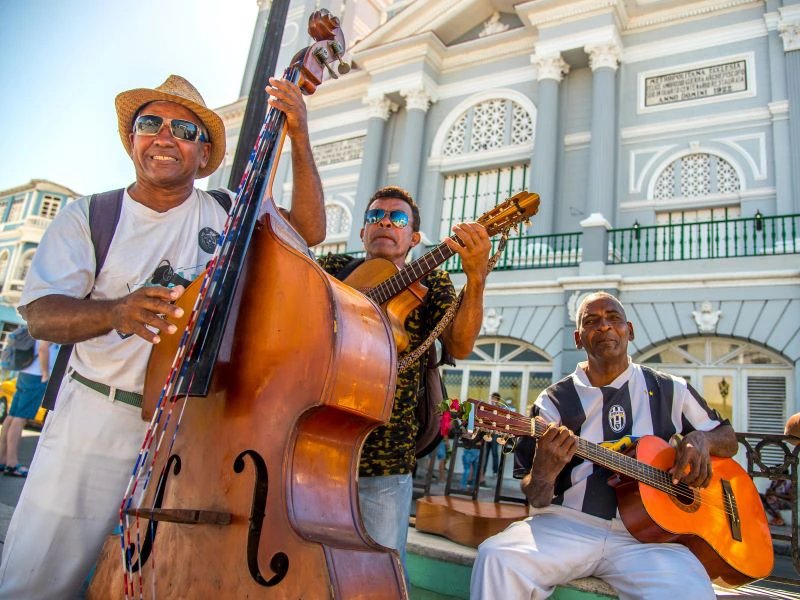Drei lächelnde Musiker spielen Kontrabass und zwei Akustikgitarren vor einem großen grauen historischen Gebäude in Santiago de Cuba, Kuba