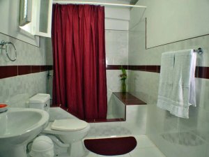 Sauberes Badezimmer mit weißen Fliesenwänden und dunkelrotem Akzentstreifen, ausgestattet mit Toilette, Waschbecken, rotem Duschvorhang und weißem Handtuch in Baracoa, Kuba