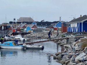 Hafen mit bunten roten und blauen Häusern an einer felsigen Küste, mehreren Booten und einer Person auf einem Holzsteg in Grönland