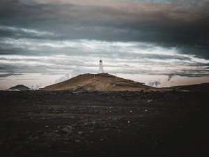 Weißer Leuchtturm steht auf einem braunen Hügel unter dunklem bewölktem Himmel mit schwarzen Lavasteinen im Vordergrund in Island