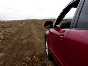 Rotes Auto fährt auf einer felsigen Schotterstraße durch eine karge Landschaft mit einem Arm aus dem Fenster in Island