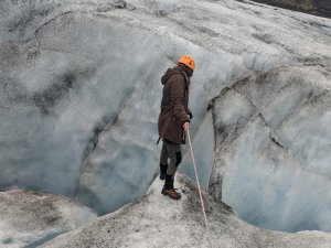 Person mit orangefarbenem Helm und brauner Jacke läuft mit einem Seil über einen blau-grauen Gletscher in Island