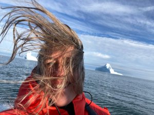 Person in roter Jacke mit wild wehenden Haaren auf dem Wasser vor Eisbergen in Grönland