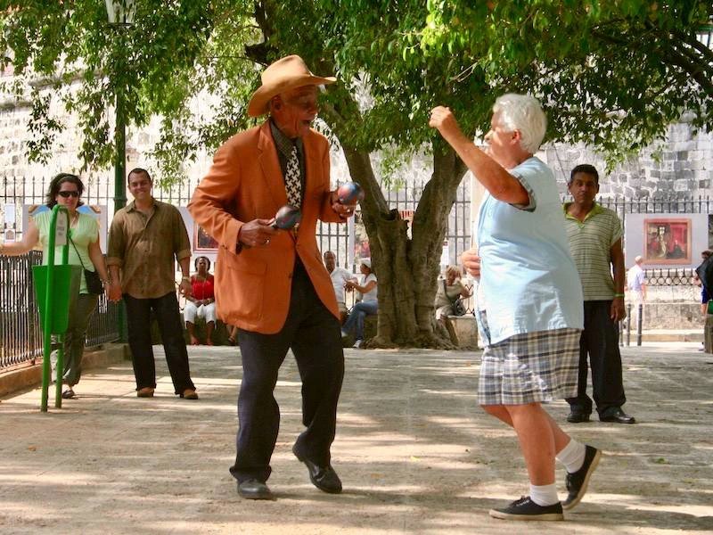 Älterer Mann mit Cowboyhut und orangefarbener Jacke schüttelt Maracas, während eine ältere Frau mit erhobenen Armen in einem sonnigen Park tanzt in Santiago de Cuba, Kuba