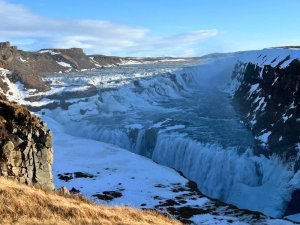 Breiter mächtiger Wasserfall fließt durch eine verschneite Schlucht unter blauem Himmel in Island