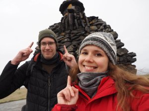 Lächelndes Paar zeigt nach oben vor einer großen dunklen Steinstatue in Island