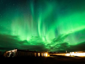 Leuchtend grünes Nordlicht zieht über einen Sternenhimmel oberhalb eines niedrigen Gebäudes mit beleuchteten Fenstern in Island