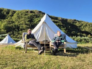 Zwei Menschen entspannen auf Stühlen vor weißen Glamping-Zelten mit grünem Hügel im Hintergrund an einem sonnigen Tag in Island
