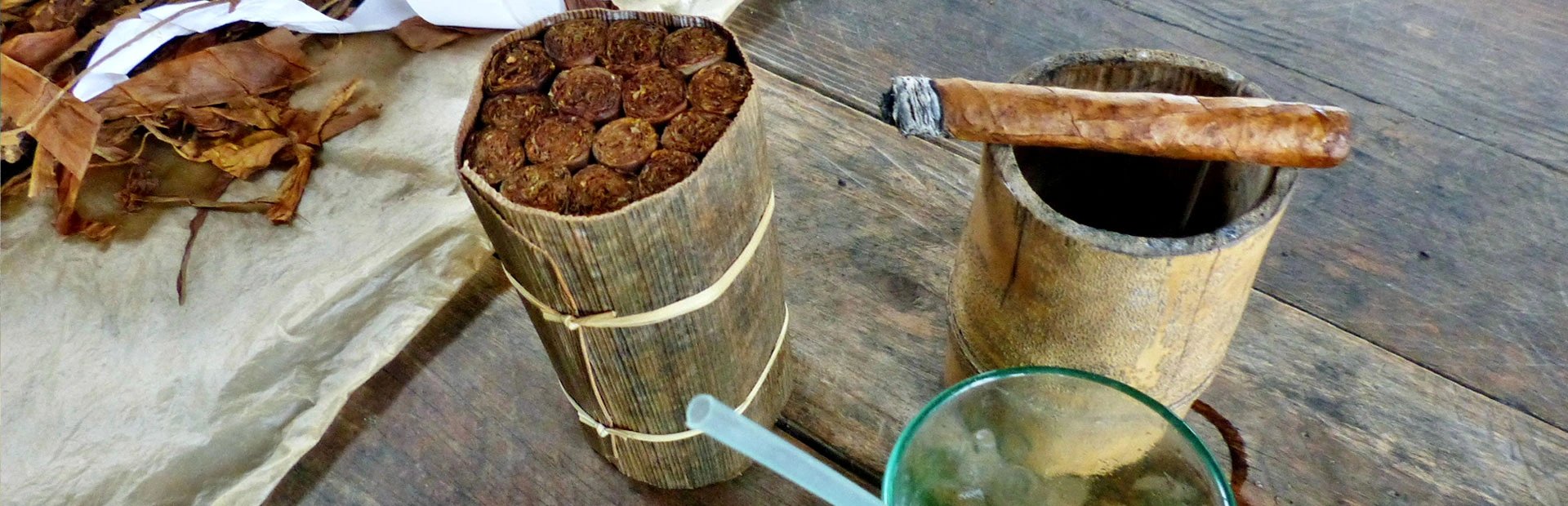 Bündel Zigarren in Blätter gewickelt, brennende Zigarre auf einem Holz-Aschenbecher und Glas mit Eis auf einem Holztisch in Viñales, Kuba
