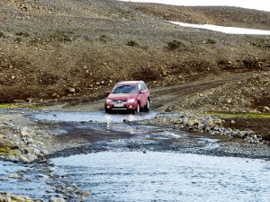 Roter SUV durchquert einen flachen Fluss in einer felsigen kargen Landschaft mit Schotterstraße und Schneeresten in Island