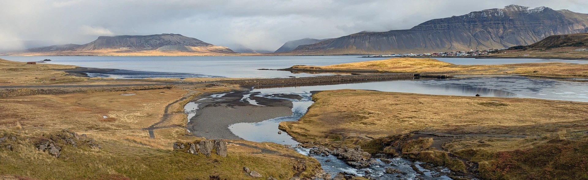 Panoramische Landschaft eines Fjords in Island mit Fluss, Dammstraße, Bergen in der Ferne und einem kleinen Dorf