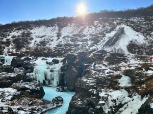 Leuchtend blauer Fluss fließt durch eine verschneite felsige Schlucht mit gefrorenen Wasserfällen und Eiszapfen unter sonnigem Himmel in Island