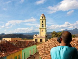 Person fotografiert einen historischen gelben Glockenturm, farbenfrohe Dächer und Berge in der Ferne unter strahlend blauem Himmel in Trinidad, Kuba