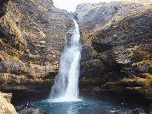 Mächtiger Wasserfall stürzt in ein blaues Becken zwischen dunklen moosbedeckten Klippen in Island