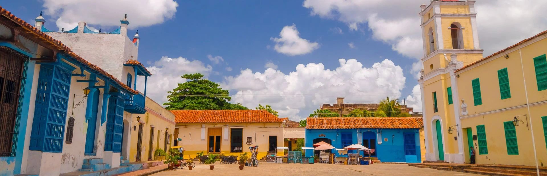 Lebhafter Stadtplatz mit farbenfrohen Kolonialgebäuden und gelbem Kirchturm unter sonnigem bewölktem Himmel in Trinidad, Kuba