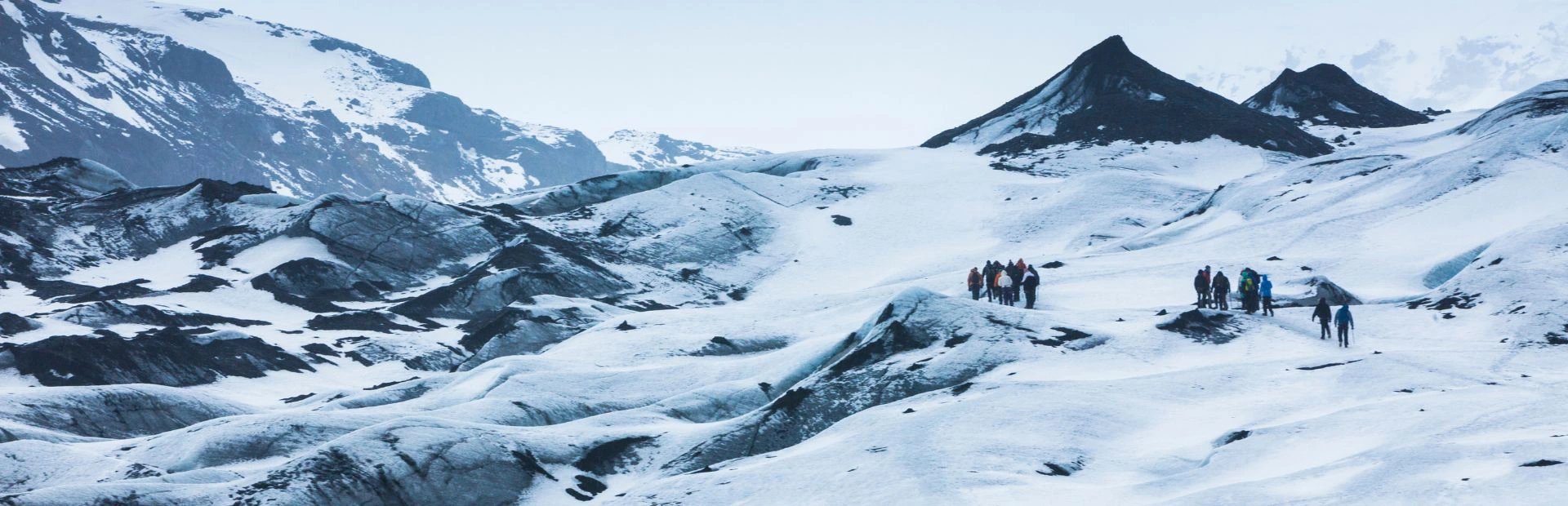 Gruppe von Wanderern überquert einen schneebedeckten Gletscher in Island mit dunklen Felsformationen und Bergen in der Ferne