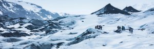 Gruppe von Wanderern überquert einen schneebedeckten Gletscher in Island mit dunklen Felsformationen und Bergen in der Ferne