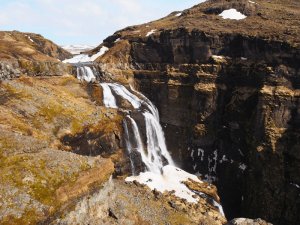 Mehrstufiger Wasserfall in einer felsigen Landschaft mit Schneeresten in Island