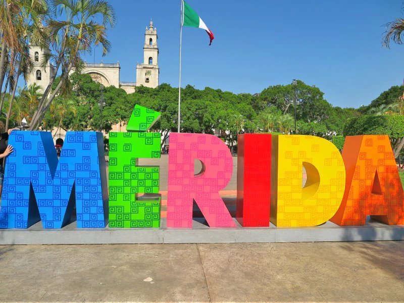 Bunte MERIDA-Buchstaben auf einem Platz mit historischer Kathedrale und mexikanischer Flagge im Hintergrund in Mérida, Mexiko