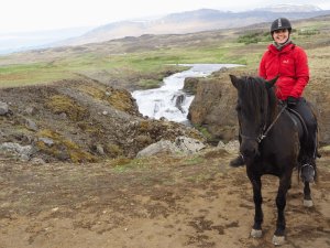 Frau auf einem dunklen Pferd mit einem Lächeln vor einem Wasserfall und rauer Landschaft in Island