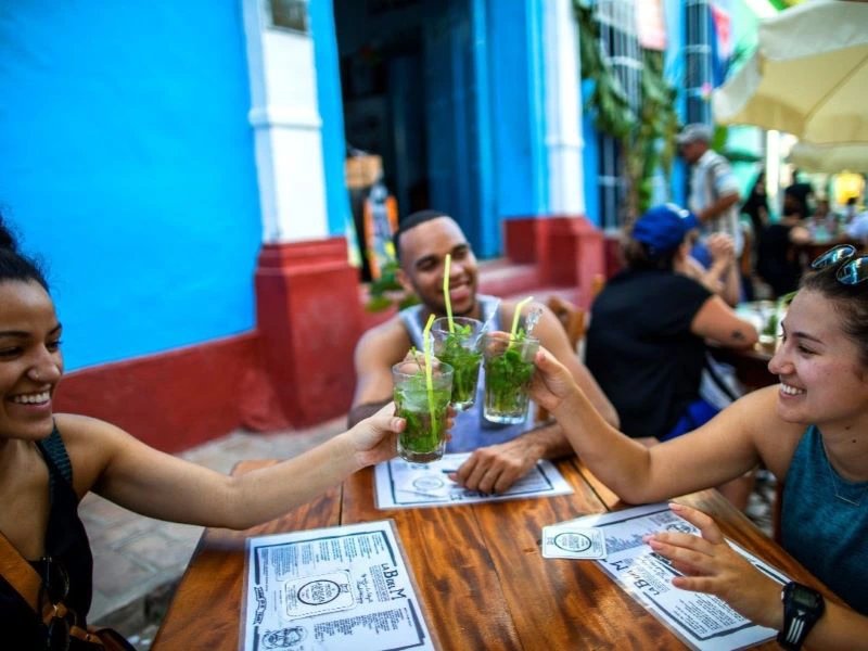 Drei lächelnde Freunde stoßen mit grünen Cocktails auf der Terrasse eines Cafés in Baracoa, Kuba