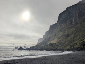 Schwarzer Sandstrand mit Wellen, einer hohen moosbedeckten Klippe und Felsnadeln im Meer unter diffuser Sonne in Island