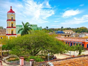 Lebhafter Stadtplatz mit gelb-rotem Glockenturm, farbenfrohen Gebäuden und großem grünen Baum unter strahlend blauem Himmel in Trinidad, Kuba