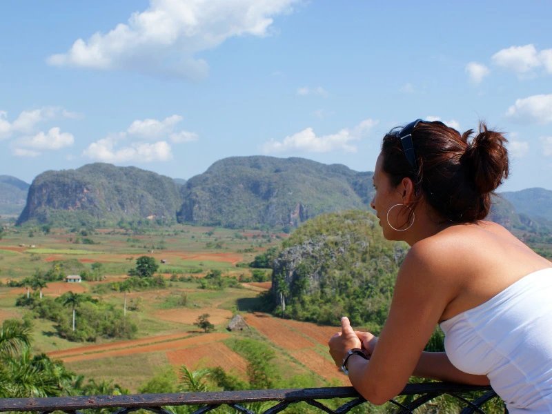 Frau in weißem Oberteil mit Blick über ein grünes Tal mit kegelförmigen Bergen und roten Feldern in Viñales, Kuba