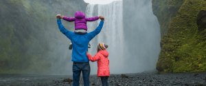 Familie mit drei Personen und einem Kind auf den Schultern blickt auf einen großen Wasserfall in Island