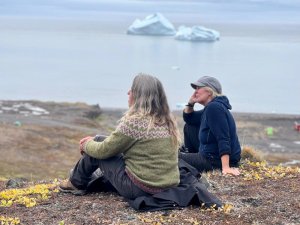 Zwei Frauen sitzen auf einem kahlen Hügel und blicken auf Eisberge in einer grauen Bucht in Grönland