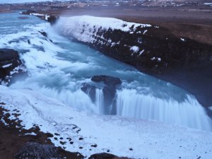 Breiter mehrstufiger Wasserfall mit türkisfarbenem Wasser fließt durch eine schneebedeckte felsige Landschaft in Island unter bewölktem Himmel
