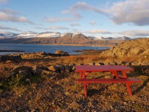 Roter Picknicktisch an einer felsigen Küste mit Blick auf eine Bucht und schneebedeckte Berge in Island