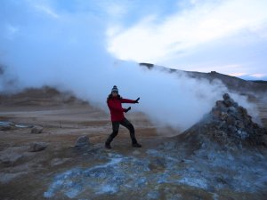 Person in roter Jacke posiert spielerisch neben einer dampfenden geothermischen Quelle in einer kargen Landschaft in Island