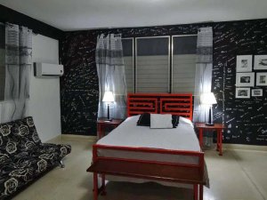 Schlafzimmer mit schwarzen Wänden voller weißer Schriftzüge, orangefarbenem Bett mit geometrischem Kopfteil, schwarz-weißem Blumensofa und gerahmten Fotos in Havanna, Kuba