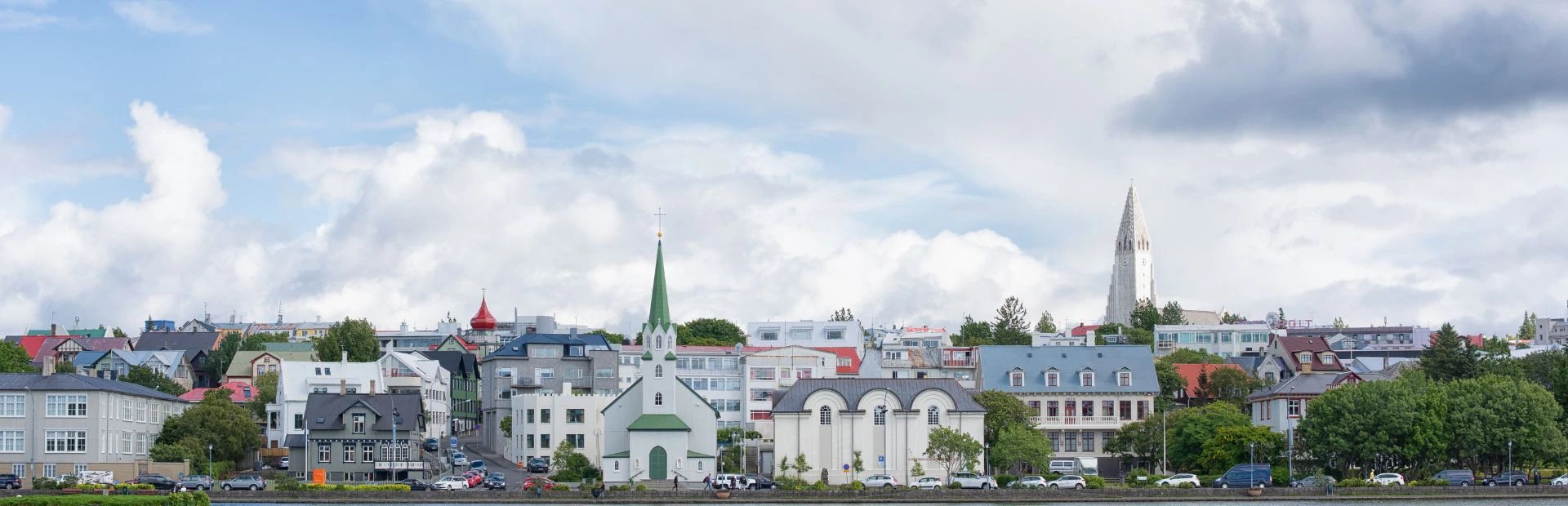 Panoramablick auf Reykjavík am Wasser mit bunten Gebäuden, einer Kirche mit grünem Turm und der Hallgrímskirkja unter bewölktem Himmel in Island