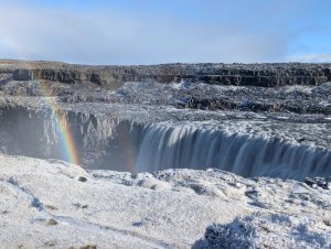 Breiter Wasserfall mit Regenbogen über dem Sprühnebel zwischen schneebedeckten Felsen in Island