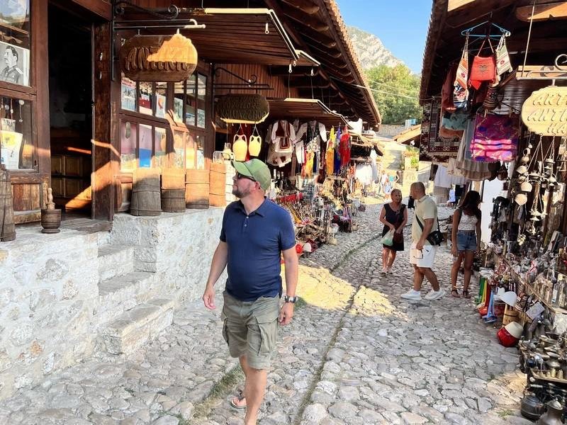 Mann geht über sonnige Marktstraße in Kruja, Albanien, mit Läden und Berg im Hintergrund