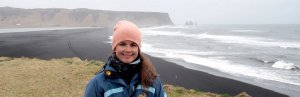 Lächelnde Frau mit rosa Mütze steht an einem schwarzen Sandstrand mit Wellen und Felsnadeln im Meer in Island