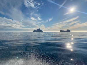 Helle Meereslandschaft mit zwei entfernten Inseln, Sonnenreflexionen auf dem Wasser und Gischt eines Boots im Vordergrund in Grönland