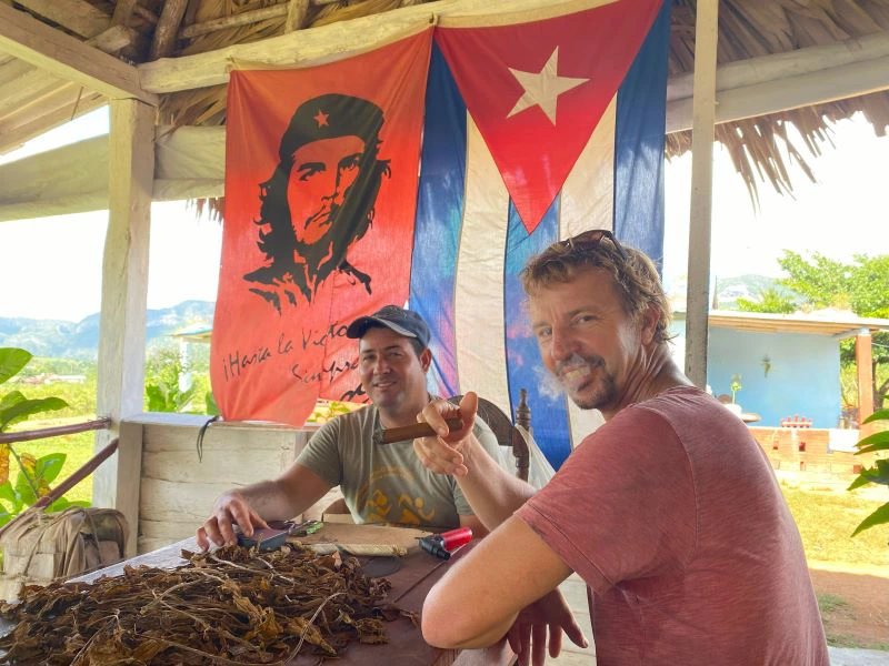 Zwei Männer an einem Tisch mit Tabakblättern, von denen einer eine Zigarre raucht, unter einer kubanischen Flagge und einem Che-Guevara-Banner in Viñales, Kuba