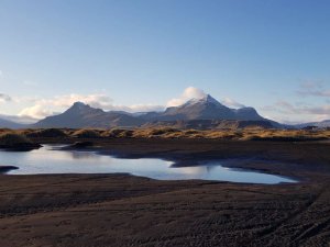 Schwarze Sandebene mit einem Wasserbecken, das den Himmel spiegelt, goldenen Dünen und schneebedeckten Bergen im Hintergrund in Island